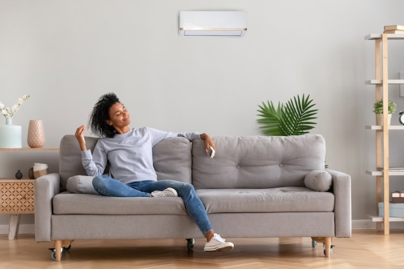 Ductless Mini Splits for Comfortable and Healthy Living. African american relaxed woman sitting on comfortable couch in living room at modern home holds air conditioner remote control enjoying breathing fresh cool air at summer or warm air at winter season (African American relaxed woman sitting on comforts.